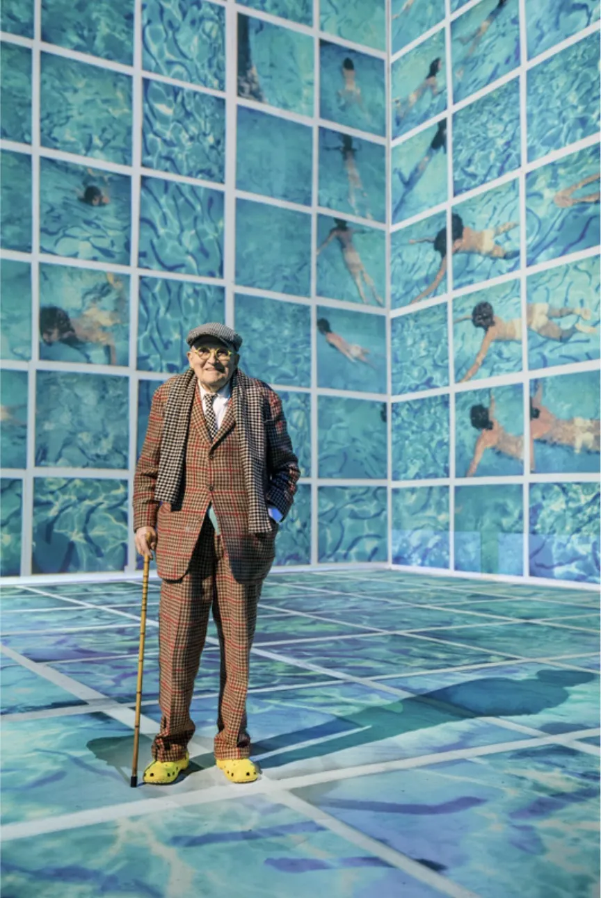 David Hockney de terno e boina xadrex, de bengala e crocs amarelos.